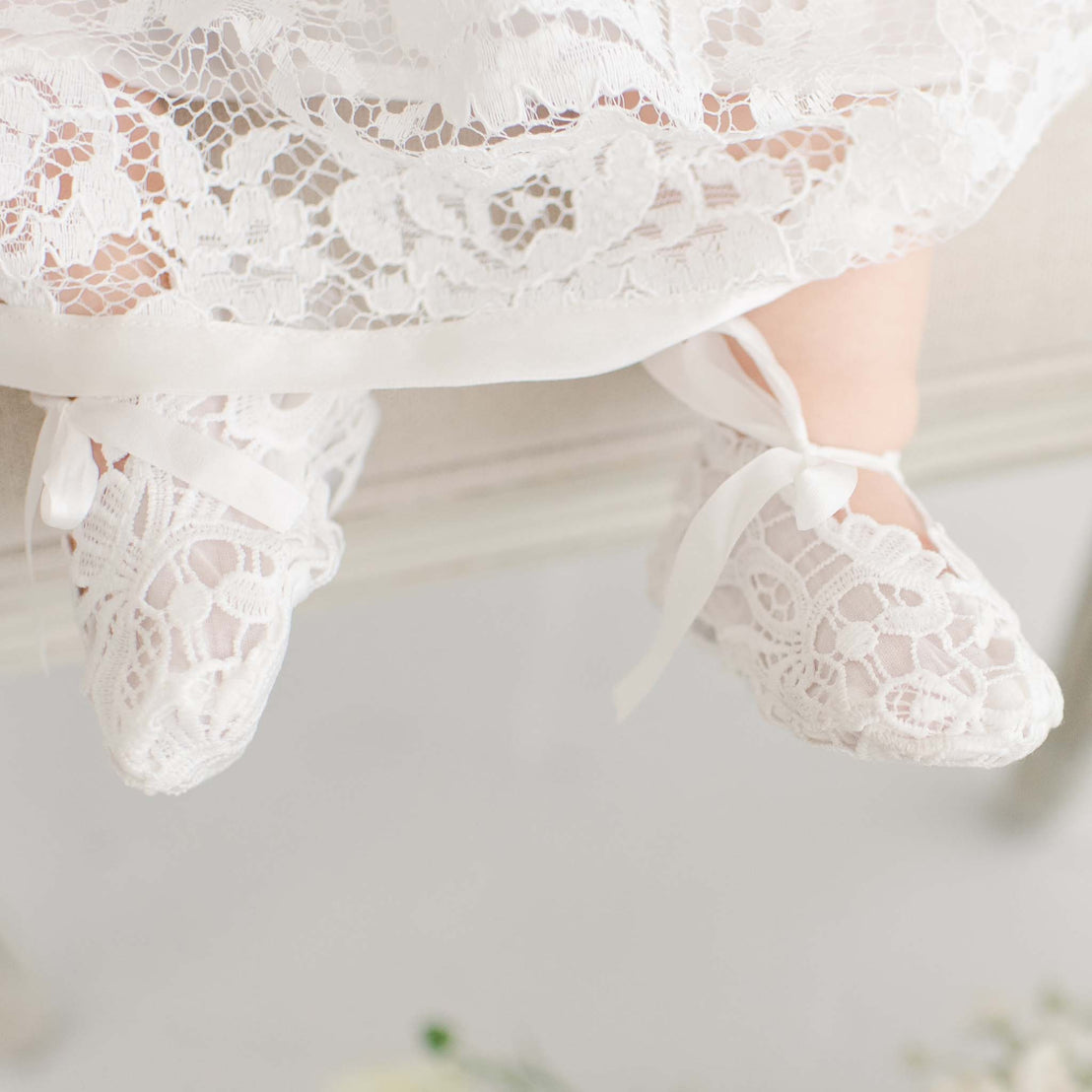 A close-up of a child's feet in delicate ivory-colored Rose Booties with silk bow ties, dangling above a soft, floral-decorated surface.