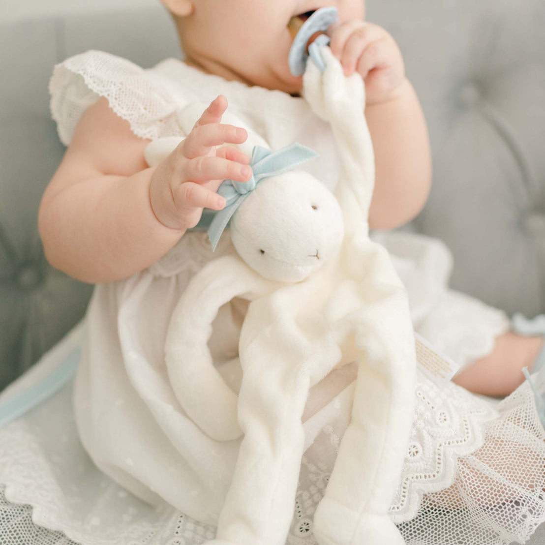 Baby girl with a sky blue Emily Silly Bunny Buddy pacifier holder. Made out of a soft white velour with a rattling head and hand loop to tie on to a pacifier.