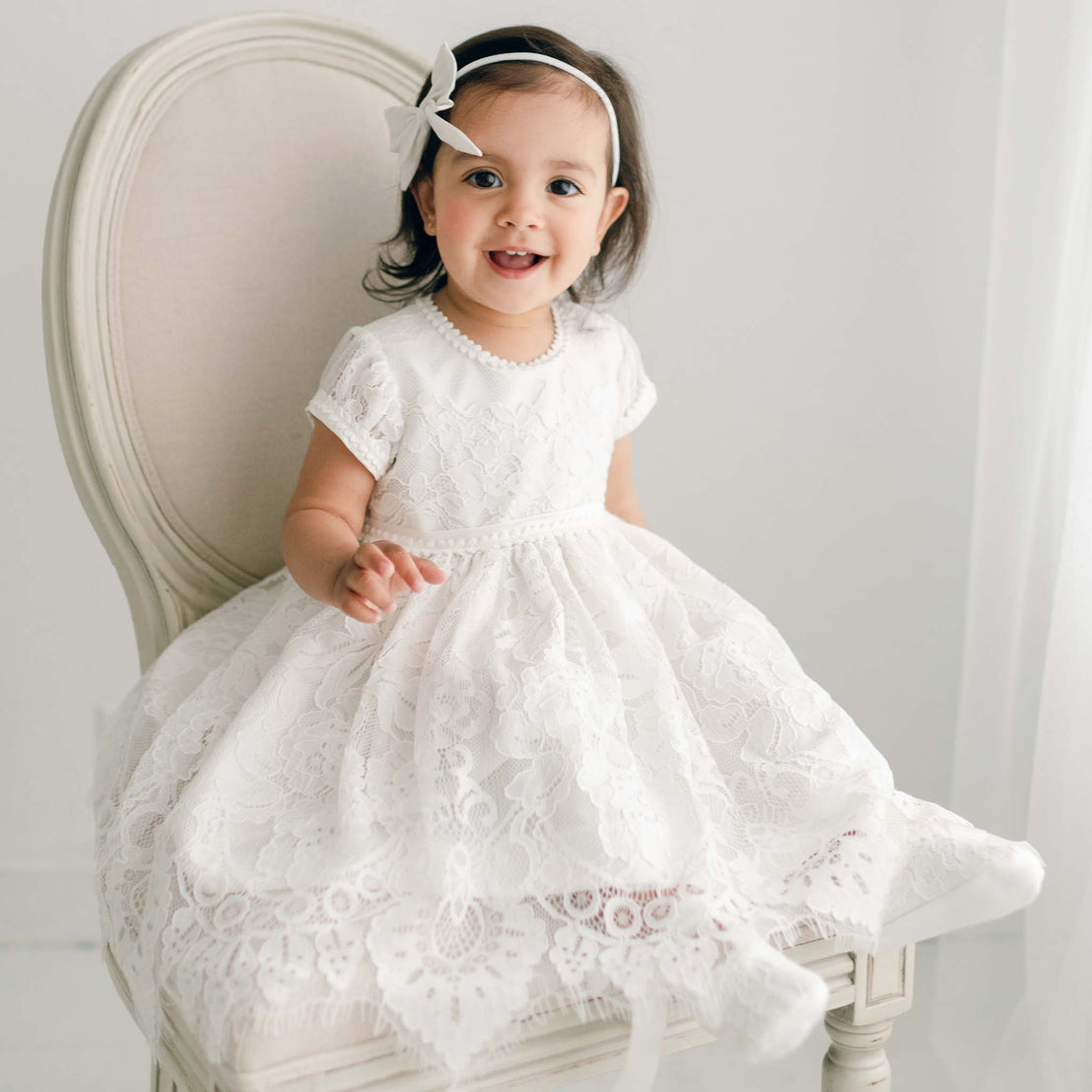 Baby girl wearing the ivory Victoria Puff Sleeve Christening Dress and Victoria Silk Bow Headband. The dress is made from an embroidered ivory lace over an ivory silk Dupioni embroidered lace.