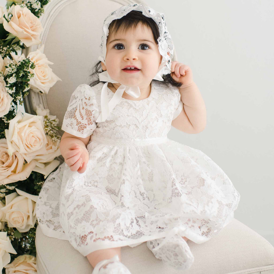 A baby girl in a white Rose Romper Dress and matching bonnet sits on a cream chair surrounded by pale pink roses, looking up with a joyful expression.