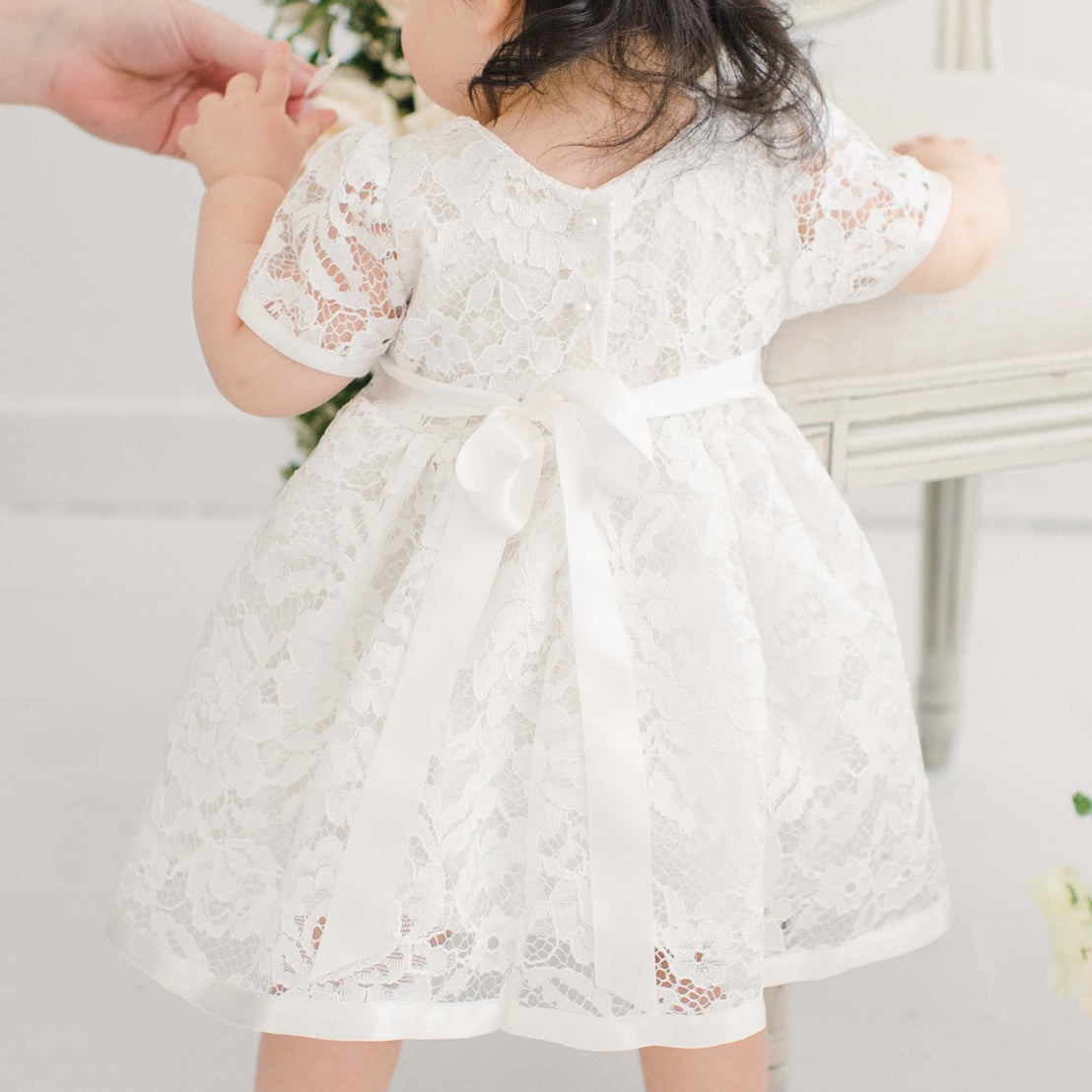 A toddler in a white Rose Romper Dress with a silk ribbon stands next to an elegant chair, reaching up towards an adult's hand, in a bright, softly lit room.
