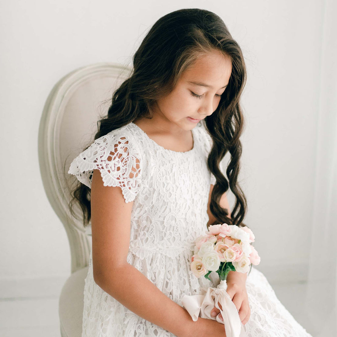 An adolescent girl wearing the Lola Girls Lace Dress. Made with cotton lining in light ivory and adorned with an all-over embroidered lace, plus a lace bodice, lacey cap sleeves, and buttons in back.