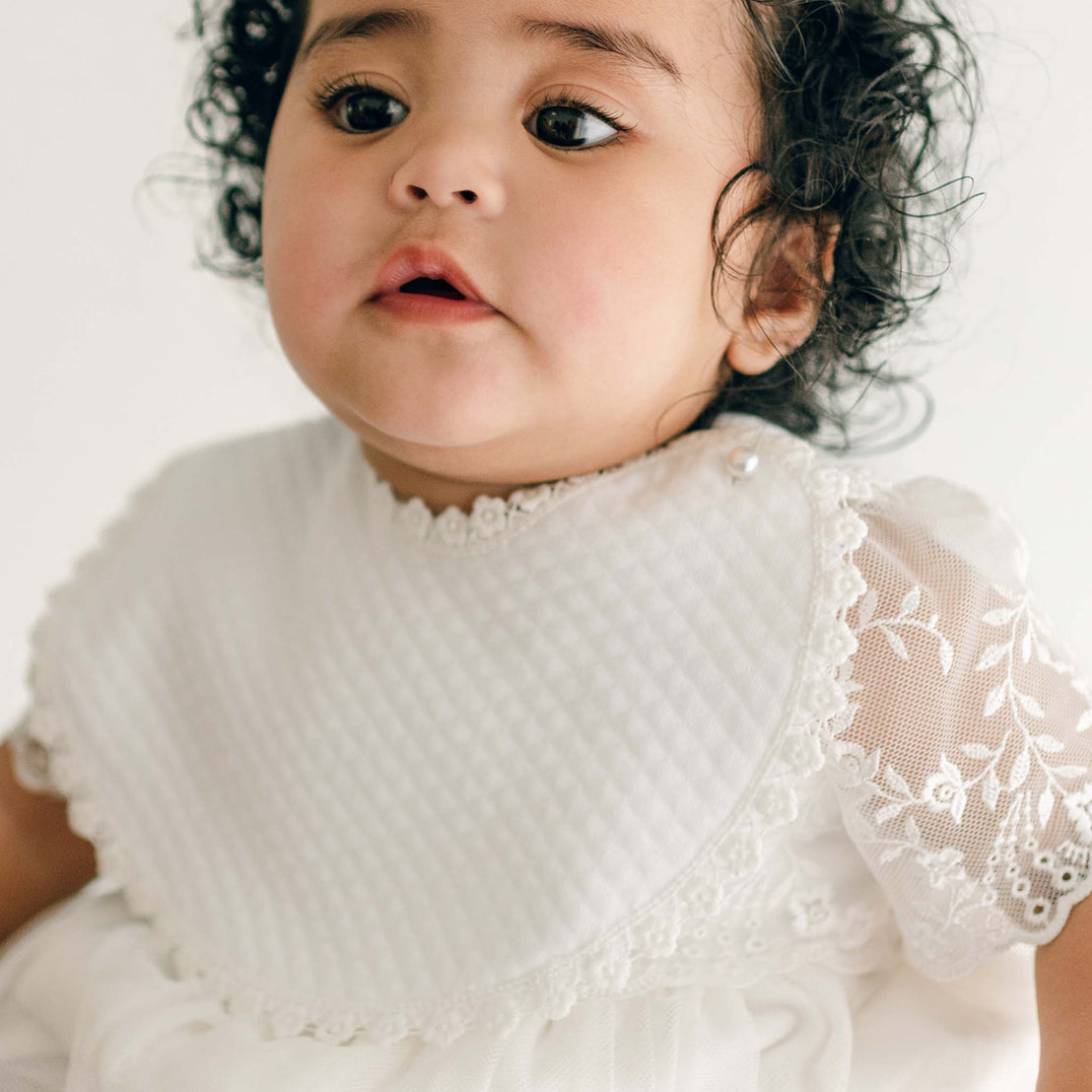 A young child with curly hair wearing a white lace special occasion dress is shown in this close-up image. The child has an Ella Bib with a quilted pattern and lace trim, and is looking slightly upwards. The background is plain and light-colored.