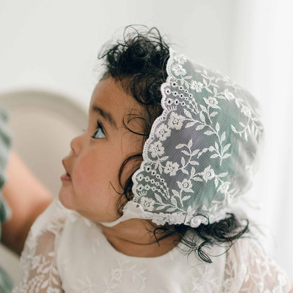 A baby is looking to the side while wearing an Ella Lace Bonnet with silk ribbon ties. The Ella Lace Bonnet features floral embroidered netting lace. The baby is dressed in a white outfit with a similar lace design, blending seamlessly with the bonnet.