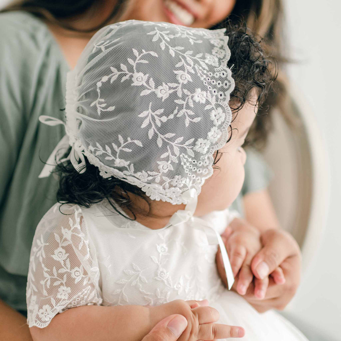 A baby wearing an Ella Lace Bonnet and dress is held by an adult dressed in a green top. The baby faces away from the camera, showing intricate details of the floral embroidered netting lace on the bonnet and gown. The adult's face is partially visible in the background, smiling.