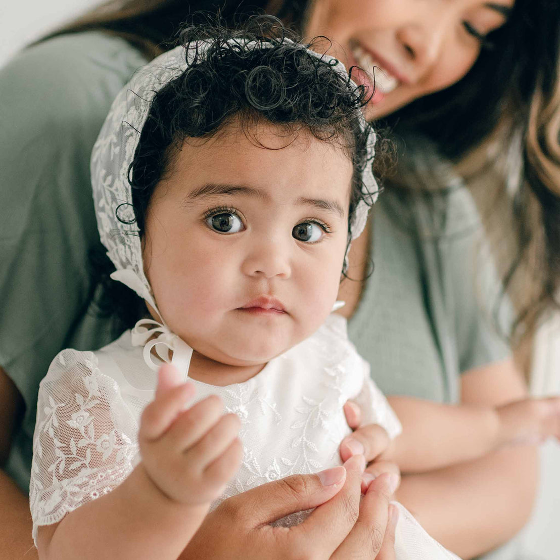 A baby with dark curly hair wearing an Ella Lace Bonnet and dress with silk ribbon ties is being held by an adult. The baby looks directly at the camera with a neutral expression, while the adult smiles in the background.
