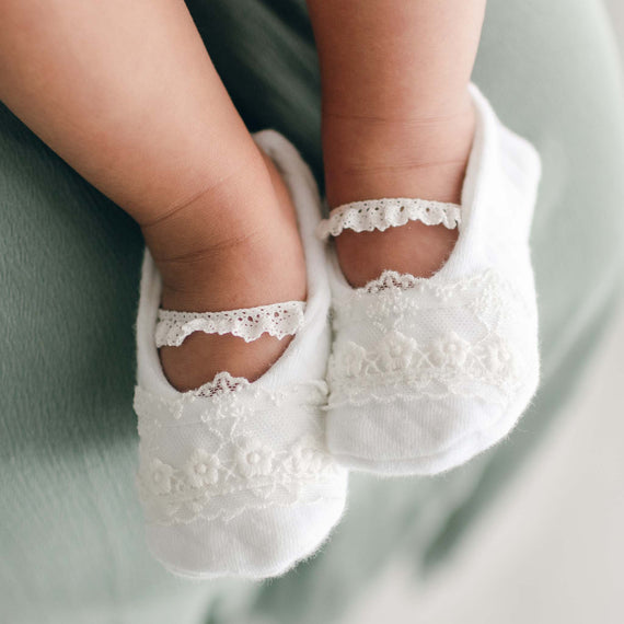 Close-up of a baby's feet wearing white lace-trimmed booties with delicate floral embroidery. The soft, green surface cradles the baby as they rest in their Ella Booties.