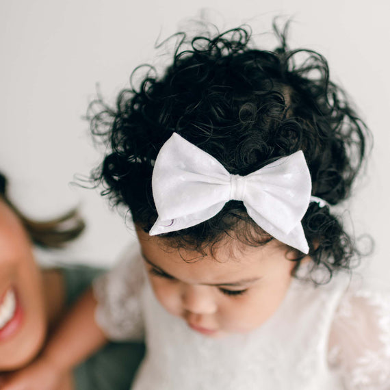 A toddler with curly hair is wearing an Ella Bow Headband on their head. They are dressed in a white outfit and looking down. Part of an adult's face is partially visible to the left. The background is plain and white.