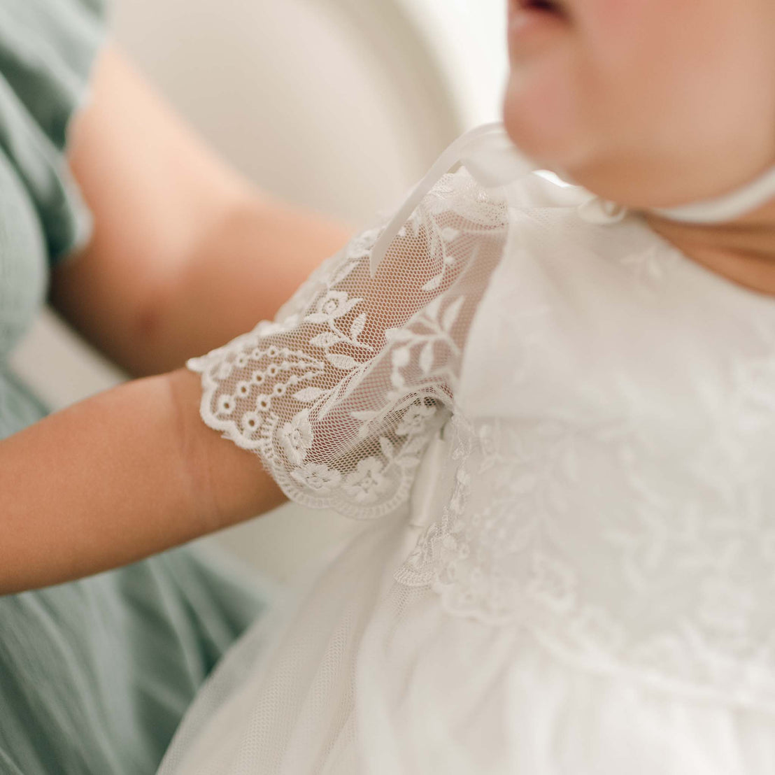 Close-up of a child wearing an Ella Christening Gown. The delicate embroidered netting lace on the sleeve is captured beautifully as the child is held by an adult dressed in green. The photo, with its softly blurred background, evokes a timeless, vintage heirloom style.
