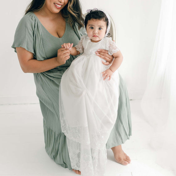 A woman in a light green dress is kneeling and holding the hands of a young child in an Ella Christening Gown. They are both looking at the camera, and the background is plain white with light sheer curtains to the side.