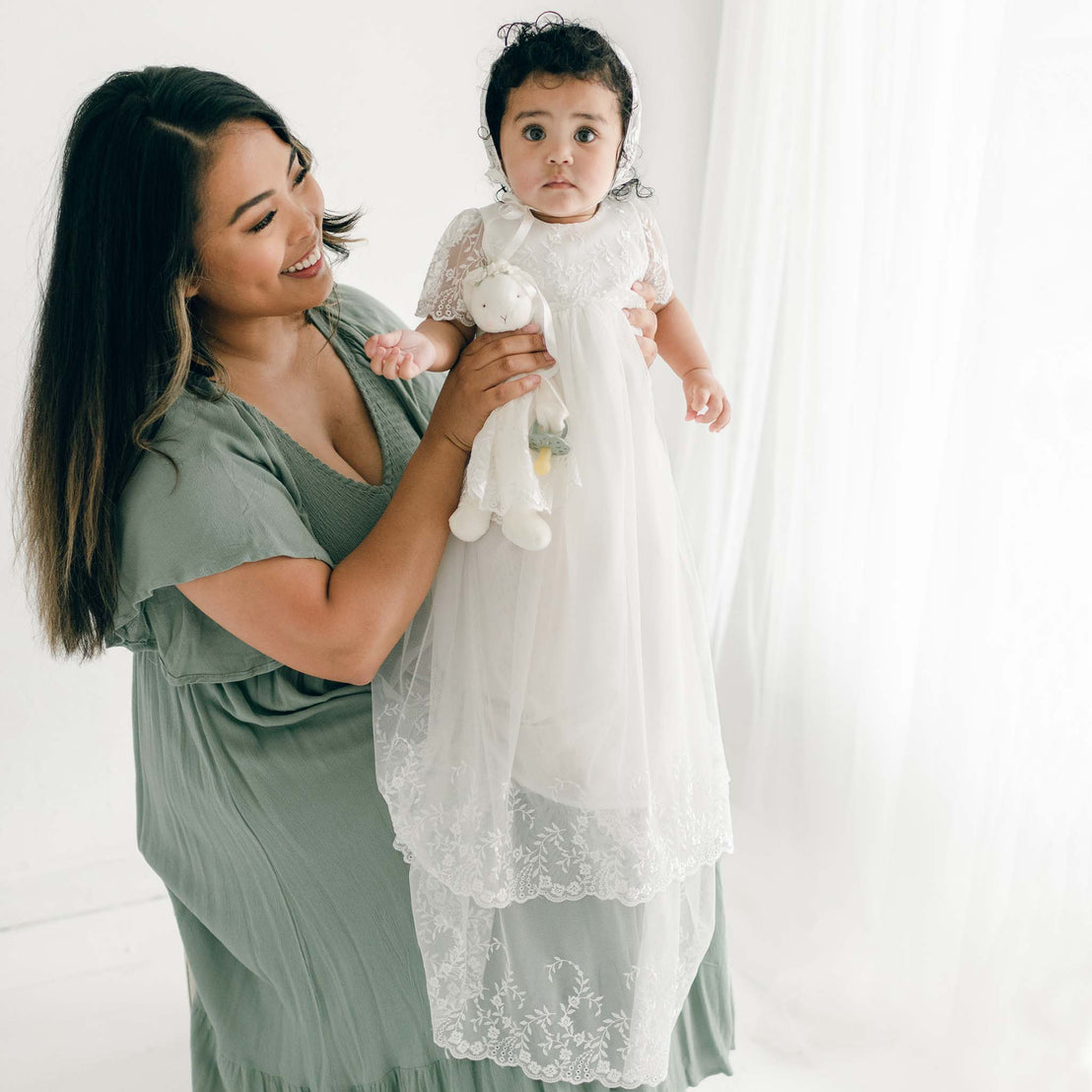 A woman in a green dress holds a baby dressed in an Ella Christening Gown, an exquisite vintage heirloom style piece adorned with embroidered netting lace. The background is minimalistic with soft lighting and white curtains.