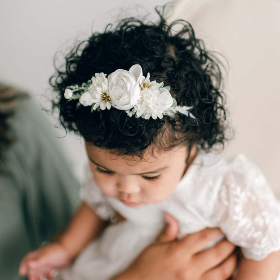 A baby with curly hair is wearing a white dress and an Ella Flower Headband adorned with white flowers and greenery while being held by an adult. The baby is looking down, and only part of the adult holding the baby is visible.