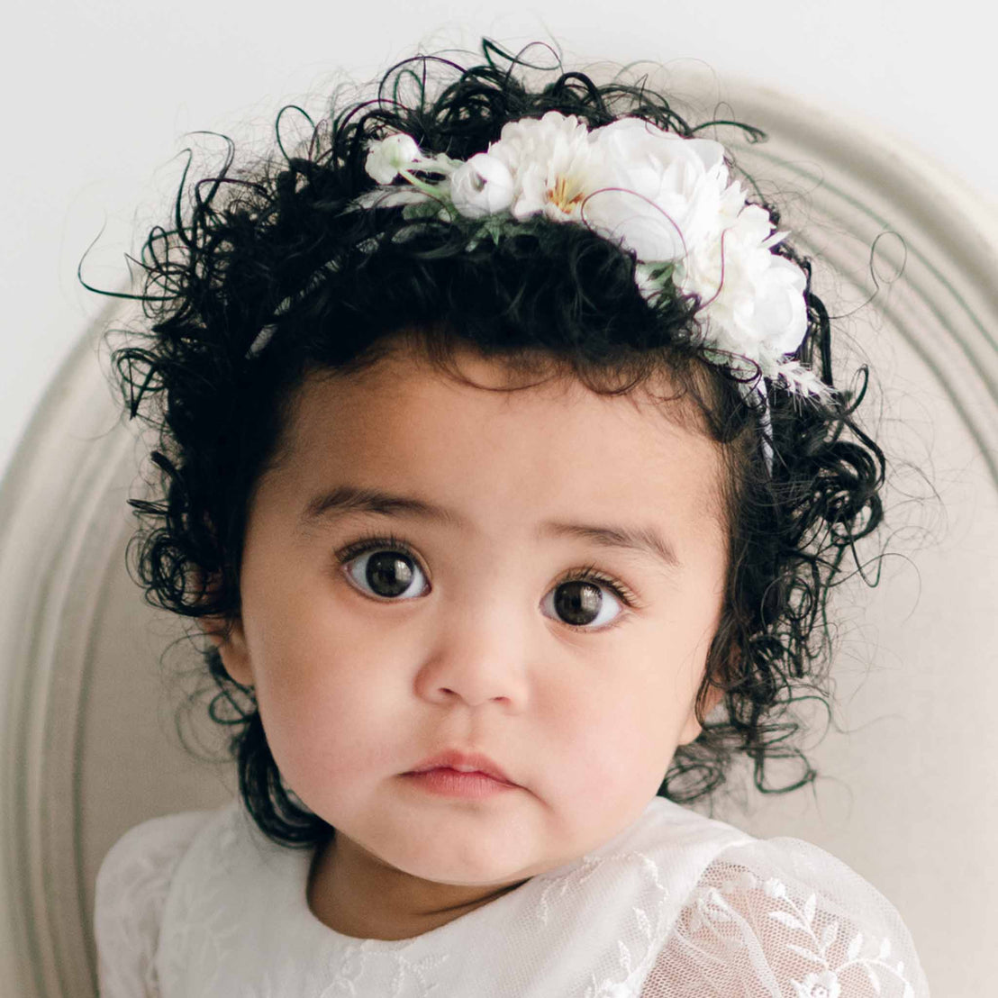 A toddler with curly black hair, adorned with an Ella Flower Headband, is seated on a beige chair. The child is wearing a white lace dress and has a neutral expression while looking directly at the camera amid the backdrop of white flowers and greenery.