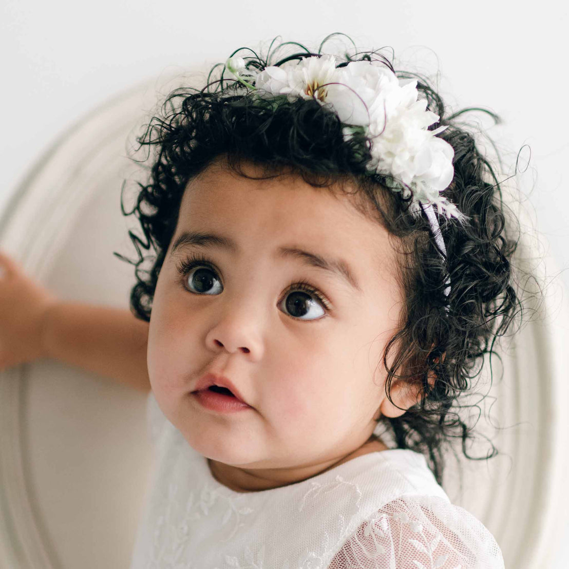 A toddler with curly hair wearing an Ella Flower Headband adorned with white flowers and greenery looks up with wide eyes. The child is dressed in a white garment, holding onto the back of a chair. The background is soft and out of focus, adding to the scene's baby comfort.