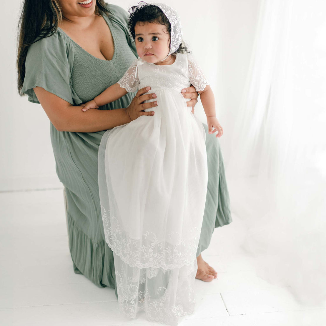 A woman in a green dress holds a baby dressed in an Ella Christening Gown with vintage heirloom style. The Ella Christening Gown, featuring embroidered netting lace, is complemented by a matching bonnet. They are standing in a minimalistic, white room with a light and airy atmosphere.