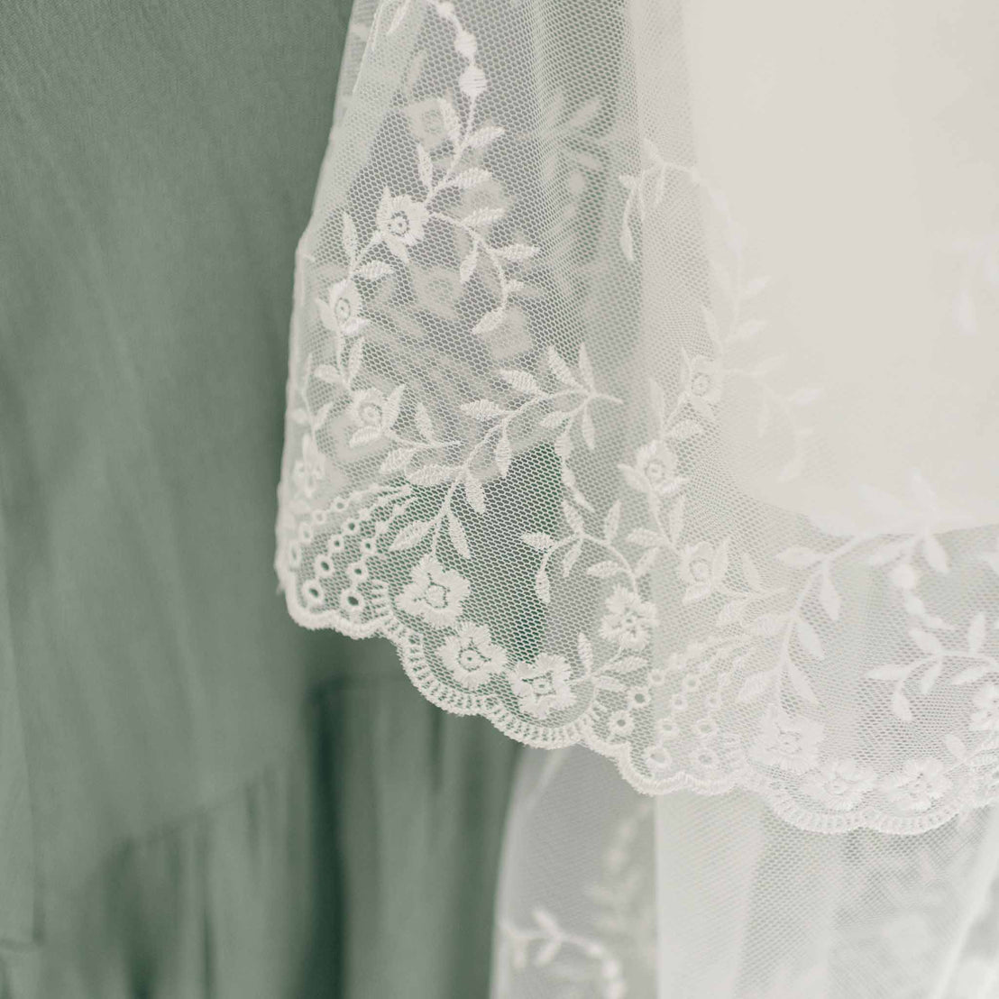 Close-up of a delicate white lace fabric with floral embroidery draped over a greenish-gray textured fabric. The vintage heirloom style lace, resembling the intricate details of an Ella Christening Gown, has a scalloped edge that creates a beautiful contrast with the smooth underlying material.