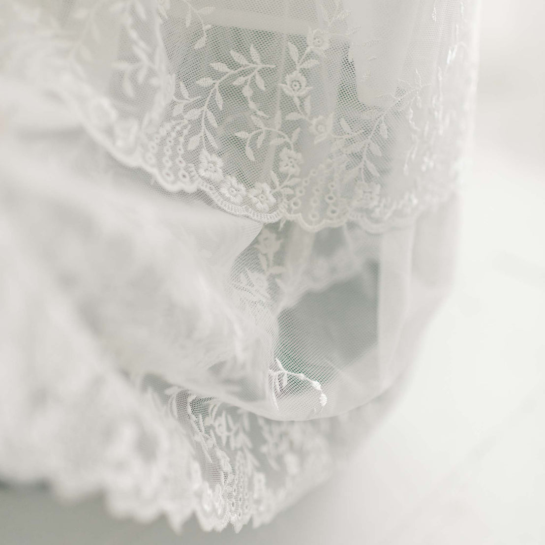 Close-up of the edge of a white lace fabric, displaying intricate floral patterns and delicate detailing. The vintage heirloom style lace, possibly part of an Ella Christening Gown, is beautifully highlighted by soft lighting that accentuates its embroidered netting lace texture and design.