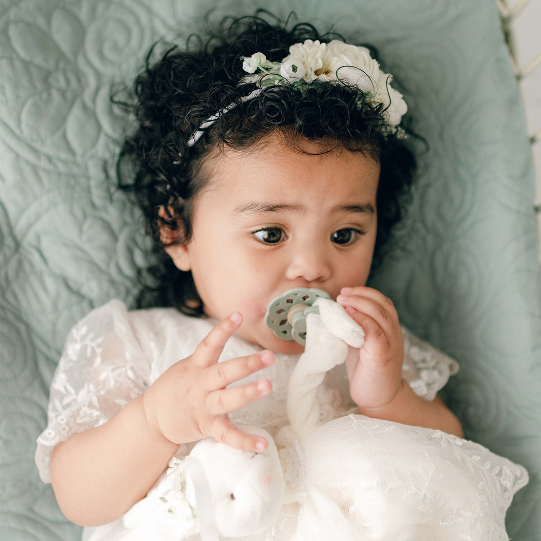 A baby with curly hair is lying on a light green quilted surface. The baby is wearing a white floral headband and a white lace dress. Holding a soft toy in one hand, the baby self-soothes using an Ella Pacifier Set | Sage & Ivory in the other, with the pacifier in their mouth.