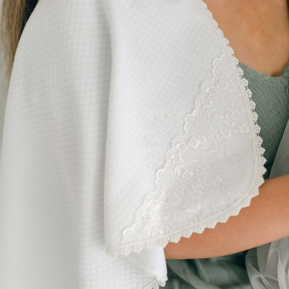 A person in a green garment holds or wears a white cloth with lace trim along the edges. The name "Lila" is embroidered into the 100% cotton fabric, showcasing exquisite personalized embroidery. The background is blurred, putting focus on the Ella Personalized Blanket's details and delicate craftsmanship.