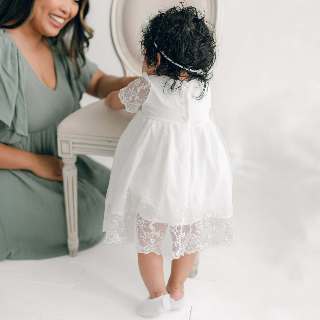 A woman in a light green dress kneels next to a toddler wearing an adorable Ella Romper Dress with embroidered netting lace. The toddler stands facing away, propping herself up on a small bench in the bright, softly-lit room with white flooring.