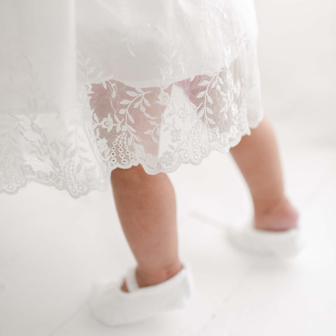 A close-up image of a baby wearing an Ella Romper Dress featuring embroidered netting lace, standing and showing only the legs and feet. The baby is also wearing white shoes with delicate patterns. The background is softly lit and neutral in color.