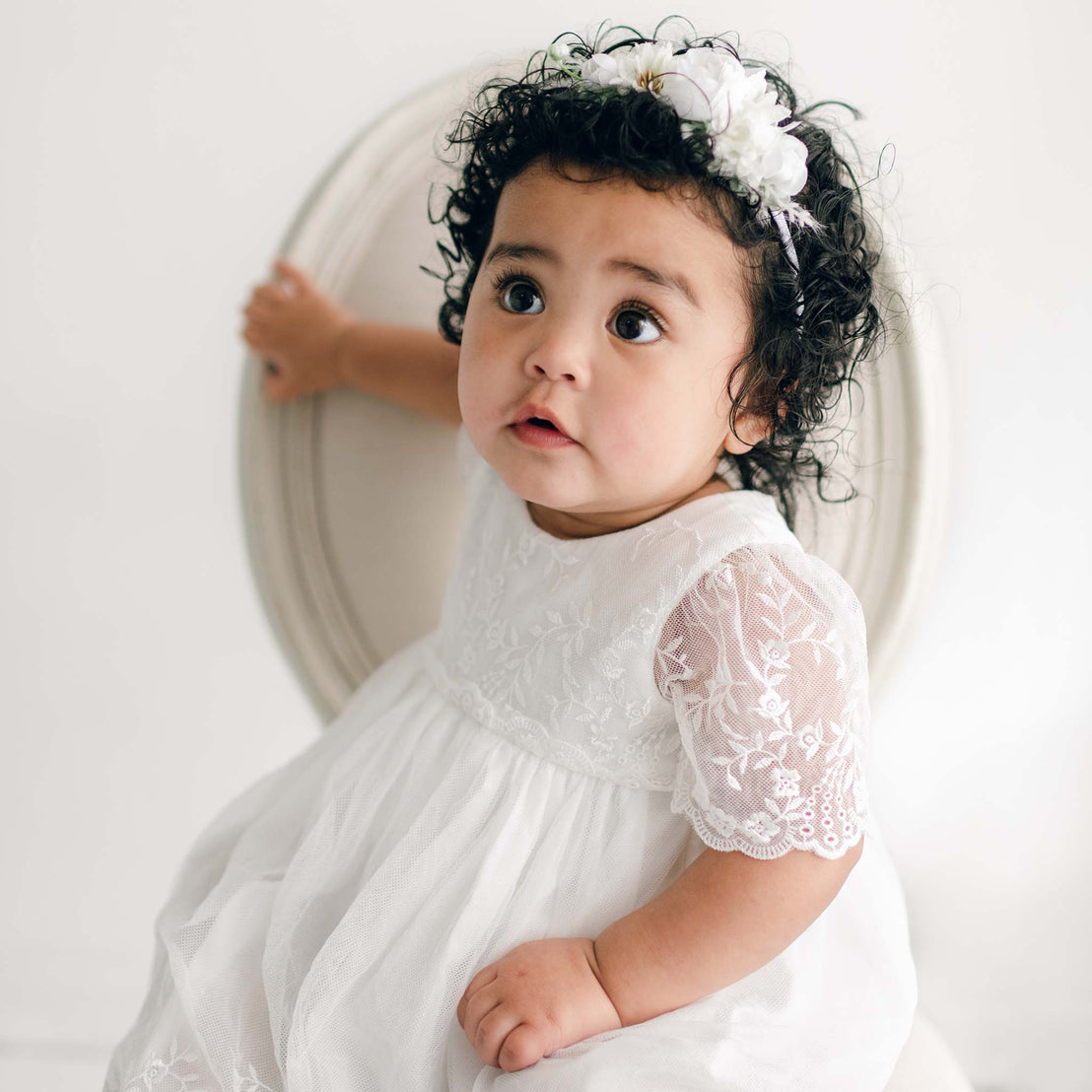 A baby with curly hair, wearing an Ella Romper Dress adorned with embroidered netting lace and a floral headband, sits on a round beige chair. She looks up with a curious expression, holding onto the back of the chair with one hand. The background is plain and light-colored.