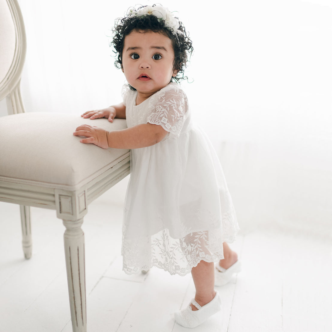 A toddler with curly hair stands next to a beige chair, wearing an Ella Romper Dress adorned with embroidered netting lace. She has a flower headband on her head and looks directly at the camera. The background is bright and airy with a white interior.