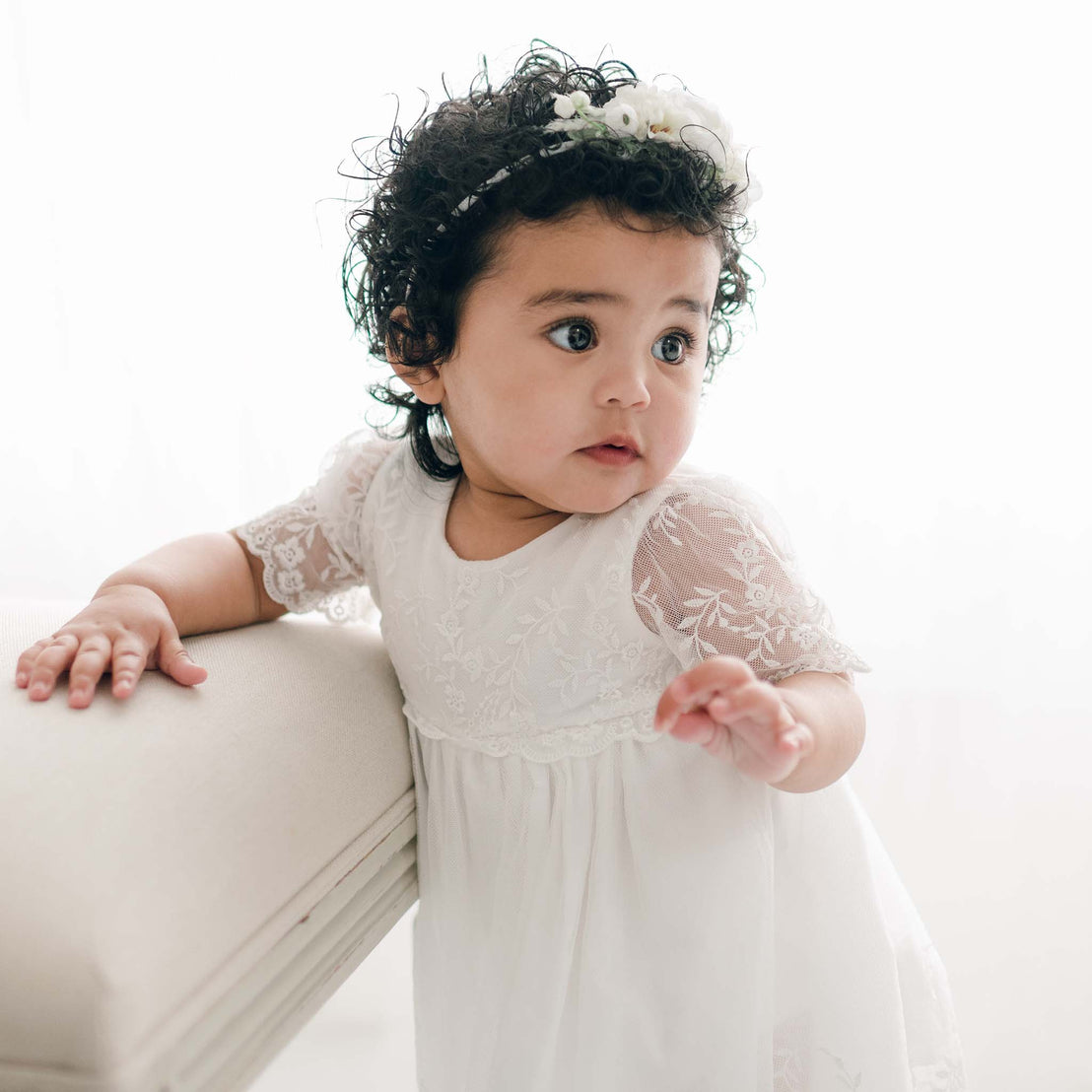 A toddler with curly hair wearing a light ivory Ella Romper Dress and a flower headband stands leaning on a cushioned chair. The background is softly lit, creating a bright, airy atmosphere. The child looks to the side with a curious expression.