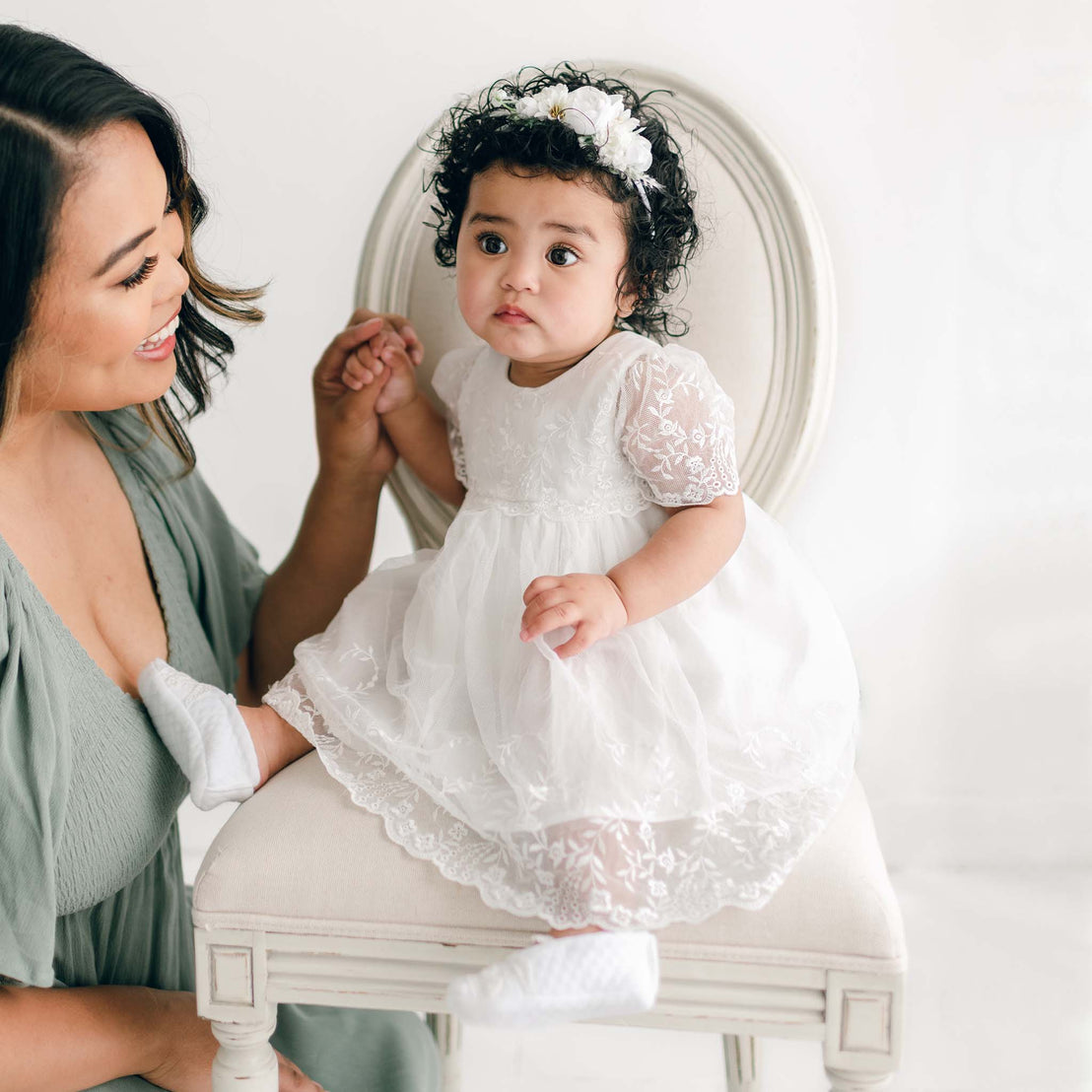 A woman in a gray dress holds the hand of a baby sitting on a chair in an adorable Ella Romper Dress, made of light ivory cotton gauze. The baby, with curly hair adorned with a floral headband, looks delightful against the light-colored indoor background.