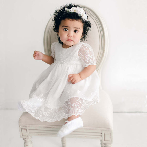 A young child with curly hair wears an Ella Romper Dress and a headband with flowers while sitting on a white, cushioned chair against a light background. The child has a curious expression and one foot resting on the edge of the chair.