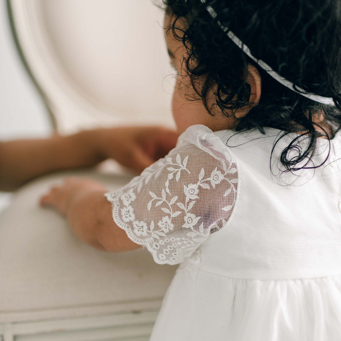 A child with curly hair, wearing an Ella Romper Dress with lace sleeves, is seen from the back. The child is touching a light-colored chair or table with both hands. The focus is on the intricate embroidered netting lace details of the dress.