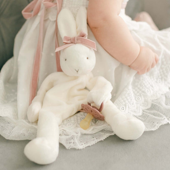 Baby girl with a blush Emily Silly Bunny Buddy pacifier holder. Made out of a soft white velour with a rattling head and hand loop to tie on to a pacifier.