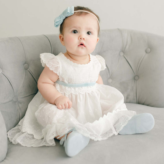 Baby girl wearing the powder blue dress Emily Dress, Bloomers, Sash Tie and Emily Velvet Bow Headband. The dress features embroidered cotton eyelet lace with a scalloped edge, lace trim and soft lace cap sleeves.