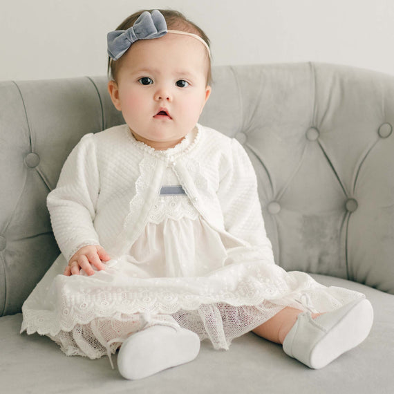 Baby girl wearing the Emily Quilted Cotton Sweater over the matching Emily Dress. The sweater is made from quilted cotton in a shade of white and is adorned with a light ivory floral lace trim and white velvet ribbon with pearl style buttons.