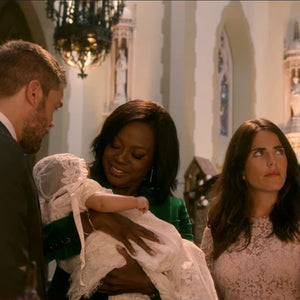 A group of four adults, two men and two women, stand in a church setting. One woman is holding a baby dressed in a white lace gown and bonnet. The background features arched windows and ornate decor.