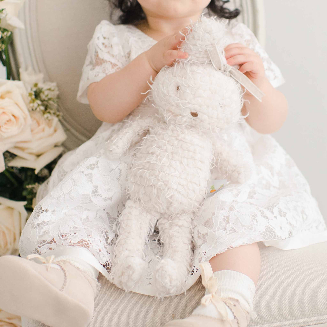 A toddler in a white lace dress holds a plush Rose Bunny while sitting on a light-colored chair, surrounded by soft pink roses. The child's face is obscured by the toy, emphasizing the Rose Bunny.