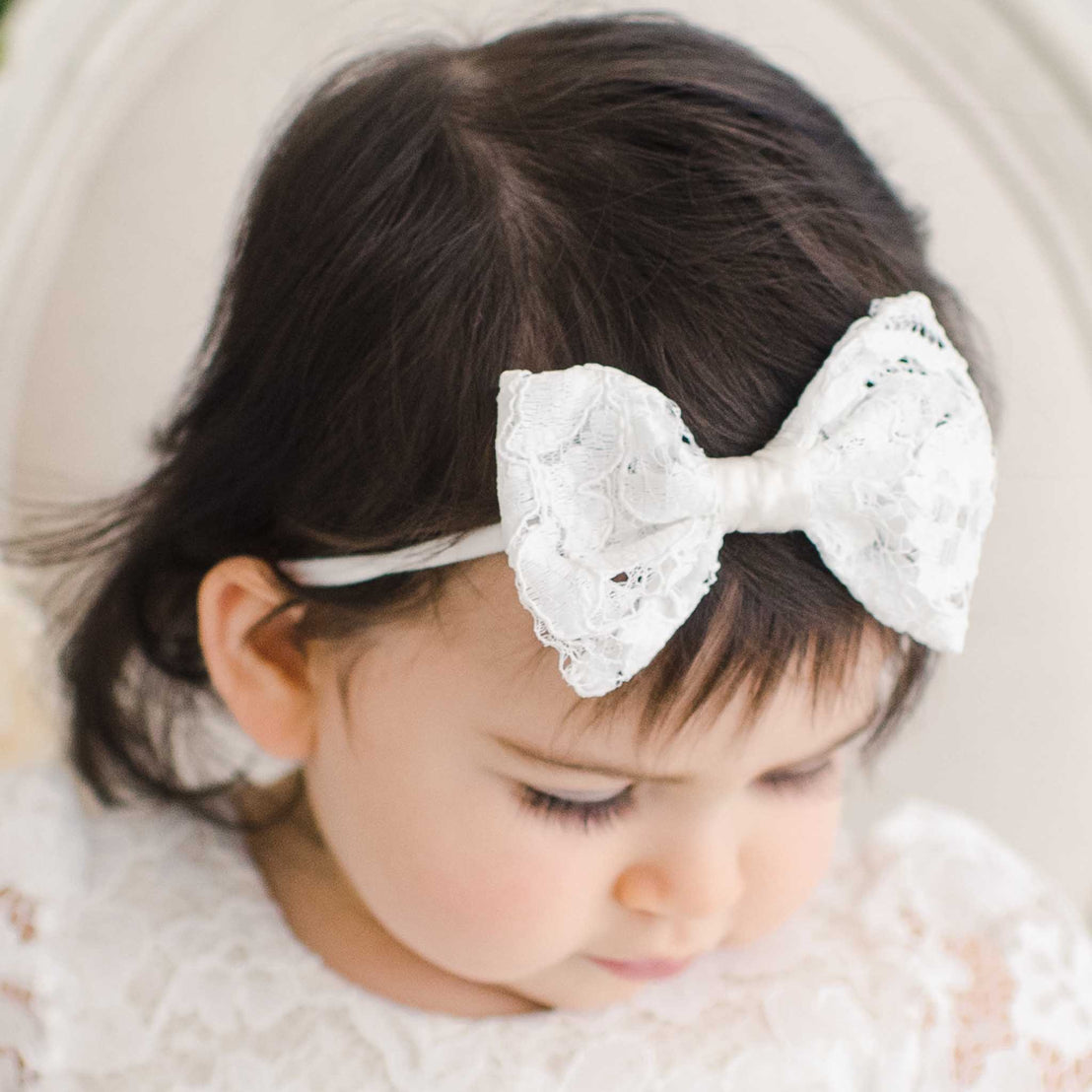 A close-up photo of a baby with dark hair wearing a large, White Rose Bow Headband, looking downward with a soft-focus background. The baby is dressed in a white lace outfit.