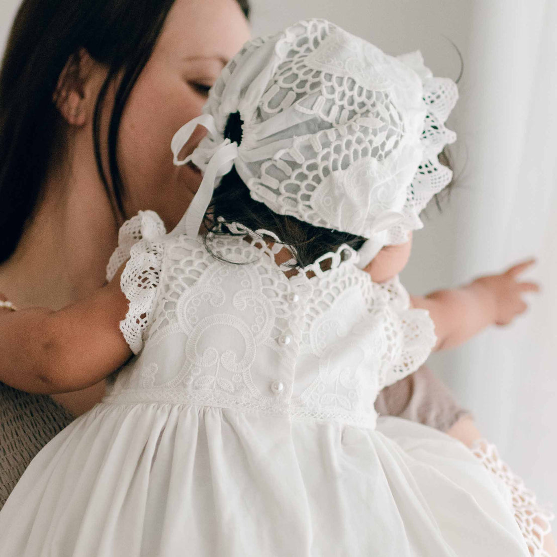 Photo of a baby wearing a Lily Christening Gown & Bonnet. She is held by her mom and the photo showcases the back of the bodice and bonnet designs