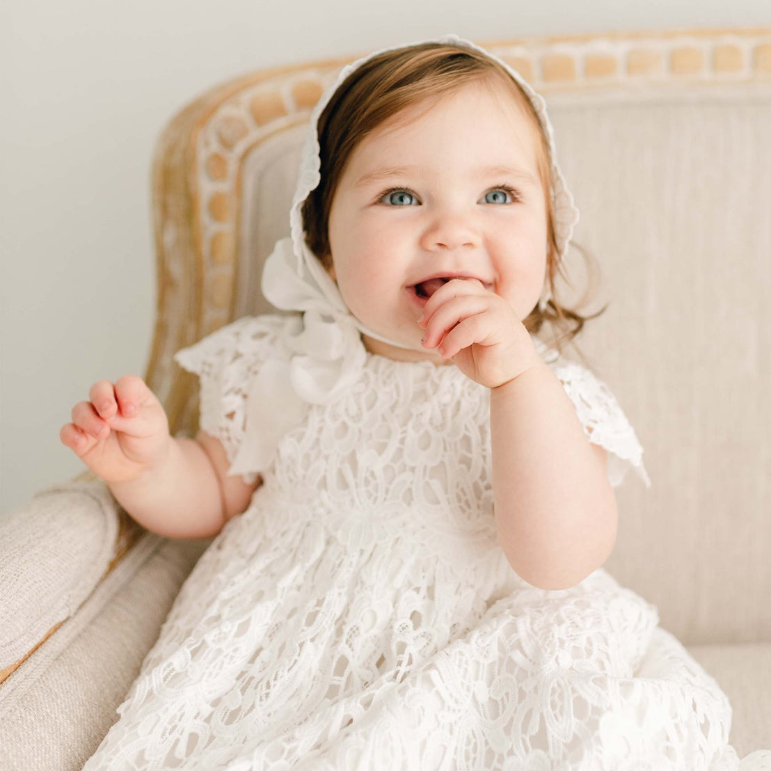 Smiling baby girl on a couch and wearing the Lola Christening Gown and Bonnet, a baptismal gown made with a cotton undergown in light ivory featuring an all-over lace overlay.