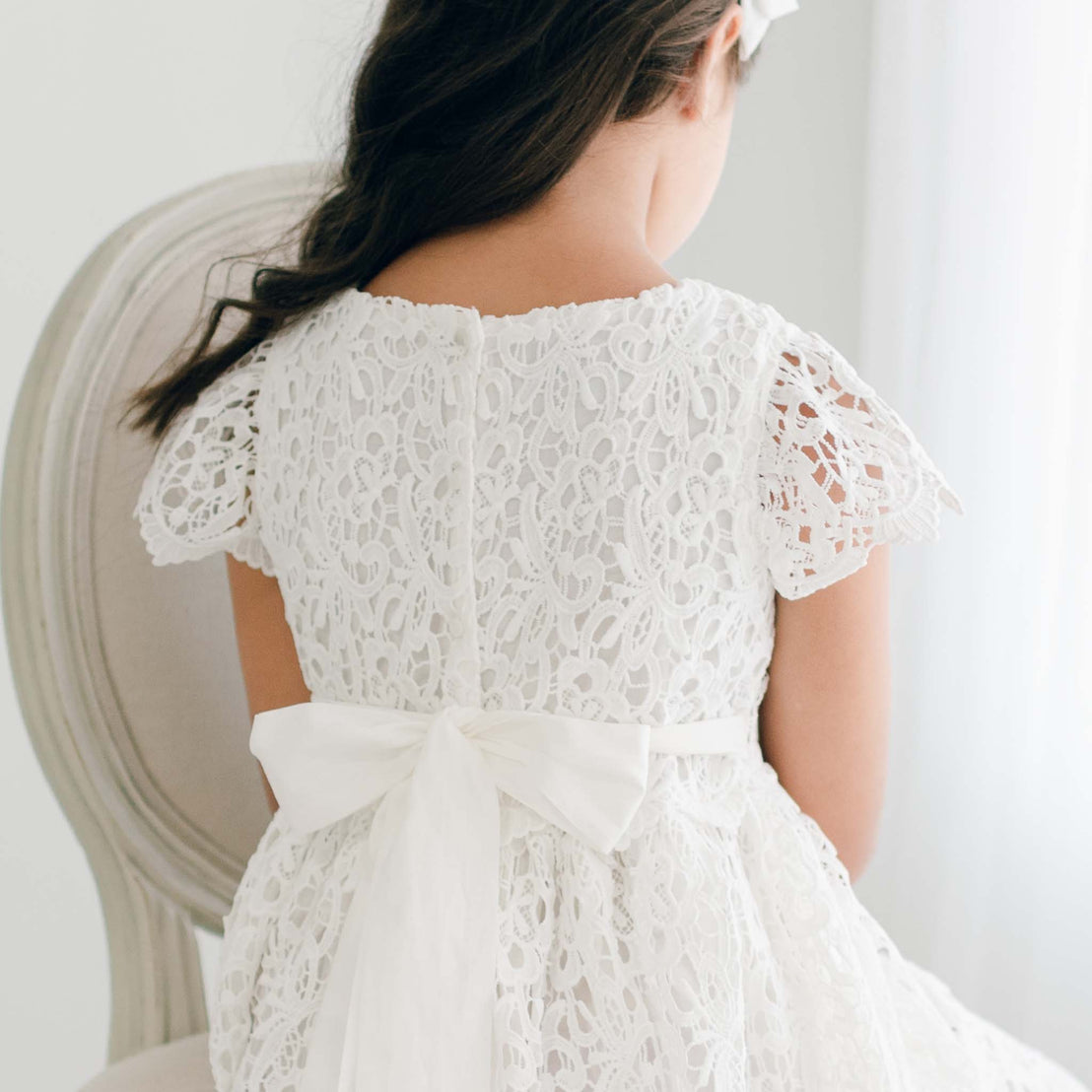 An adolescent girl wearing the Lola Girls Lace Dress. Made with cotton lining in light ivory and adorned with an all-over embroidered lace, plus a lace bodice, lacey cap sleeves, and buttons in back. Detail shows sash ties on the sides tied together.