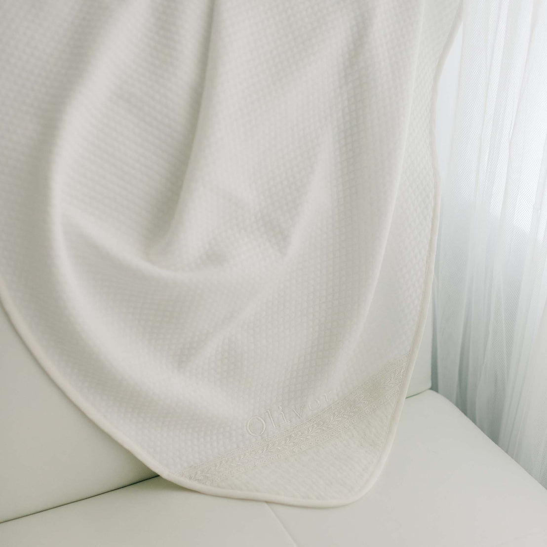 The ivory Oliver Personalized Blanket draped over the arm of a white leather sofa. The blanket is crafted from a textured pima cotton with the name "Oliver" embroidered in ivory thread on the corner of the blanket.