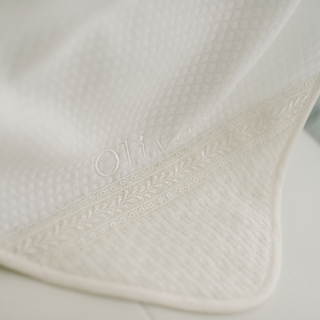 A close-up of the ivory Oliver Personalized Blanket with intricate embroidery at the edge. The handmade blanket, a personalized heirloom, features the name "Oliver" embroidered on it in ivory thread.