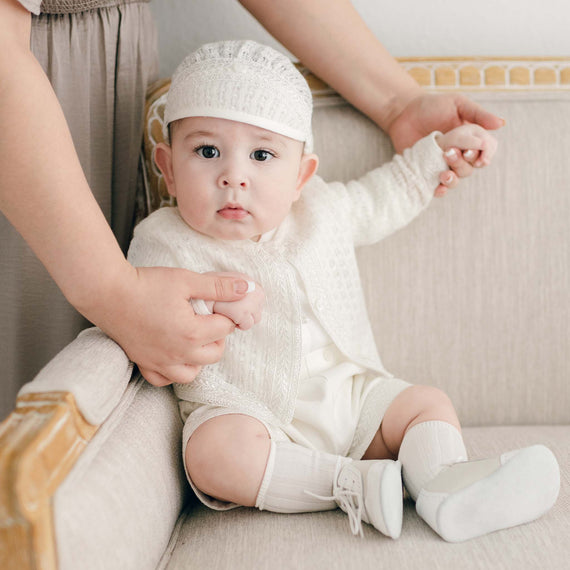 A baby boy in the light ivory Oliver Sweater Shorts Suit sits on a couch, holding a parent's hand. The baby wears the light ivory Oliver Knit Hat and looks directly at the camera with a serious expression.
