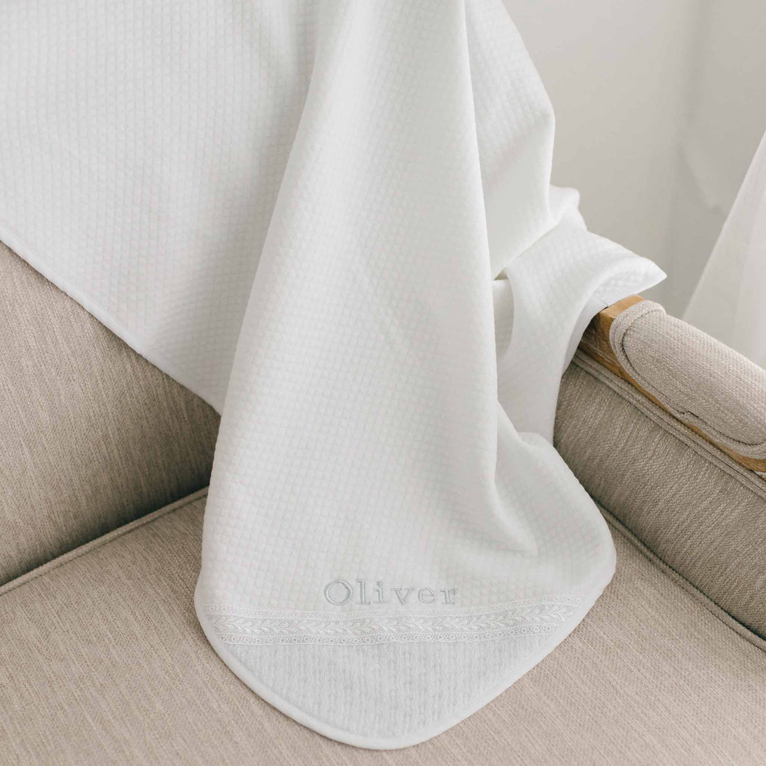 A white Oliver Personalized Blanket drapes over the armrest of a beige upholstered chair. The blanket is crafted from a white textured pima cotton and features the name "Oliver" embroidered on the corner.