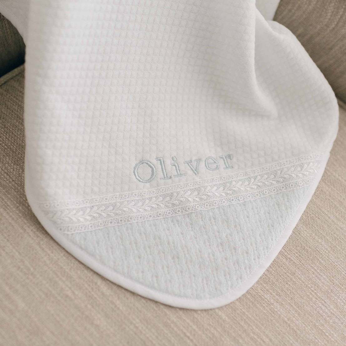 The white Oliver Personalized Blanket featuring the name "Oliver" embroidered in light blue thread is draped over a beige couch. The personalized blanket includes decorative lace trim near the embroidered name.