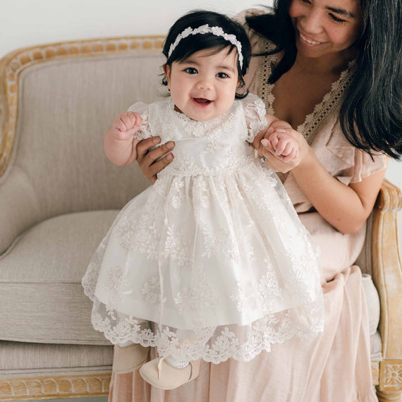 Smiling baby girl in silk and lace baptism dress, lace headband and suede shoes. Sitting on mom's lap.