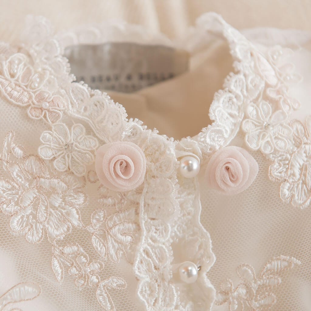 Close-up of a delicate lace garment with intricate floral embroidery. The fabric features two small pink rosettes and pearl-like buttons. The lace has a floral pattern and an elegant, vintage appearance.