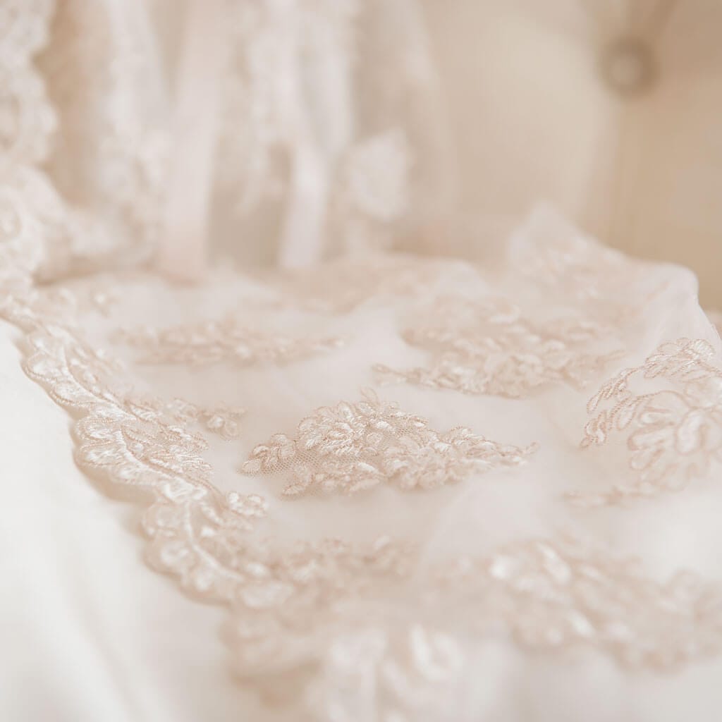 Close-up of delicate lace fabric in soft beige tones, adorned with intricate floral patterns, draped over a light, blurred background. The texture and detailing of the lace are in clear focus.
