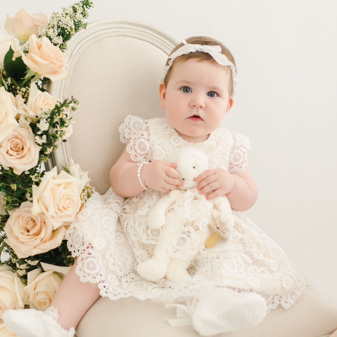 Baby girl holding plush lamb toy in her Poppy blessing dress. 