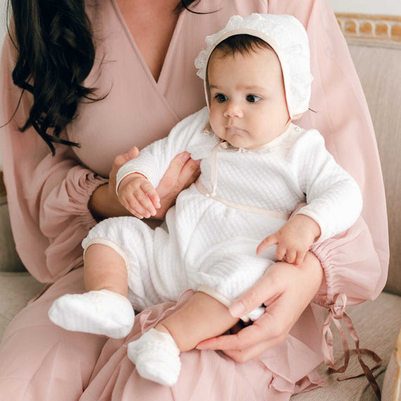 Baby girl wearing the Tessa Quilt Romper, with her mother by her side. The romper is made from a white quilted cotton and features ivory Venice Lace along the neck and sleeves and pink champagne silk trim at the cuffs, waist and neck.